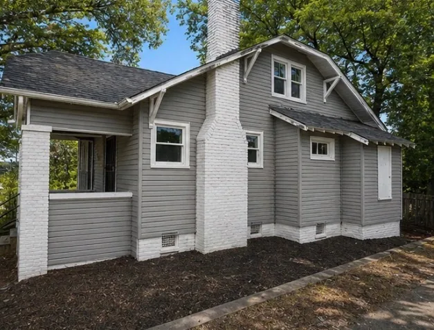 The same house after renovation, clad in grey lap siding with white trim, the chimney painted white and the original roof brackets kept