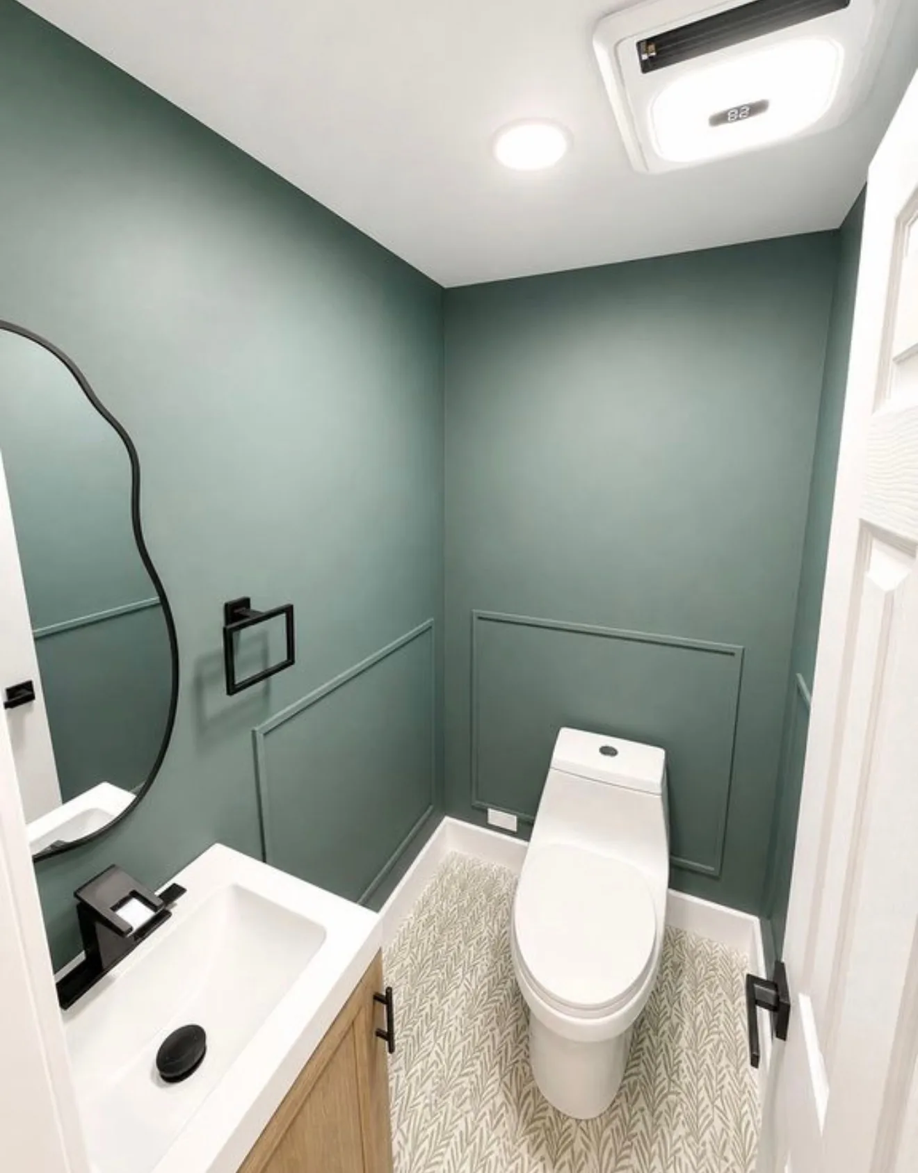 A powder room in sage green with panel moulding, an irregular black framed mirror, an oak vanity and patterned leaf tile underfoot
