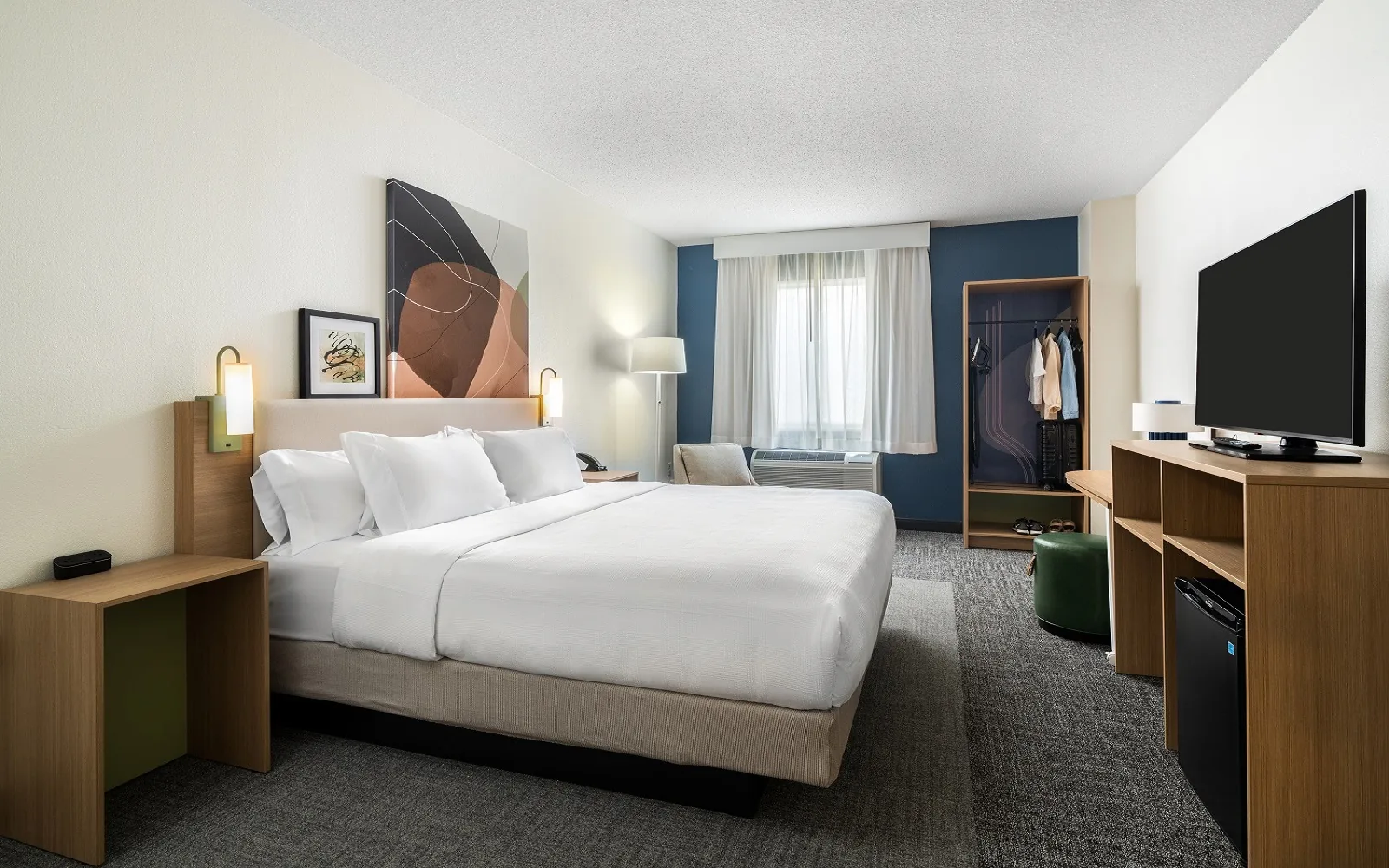 A finished king guest room at Spark by Hilton with a navy accent wall, oak casegoods and a green upholstered ottoman