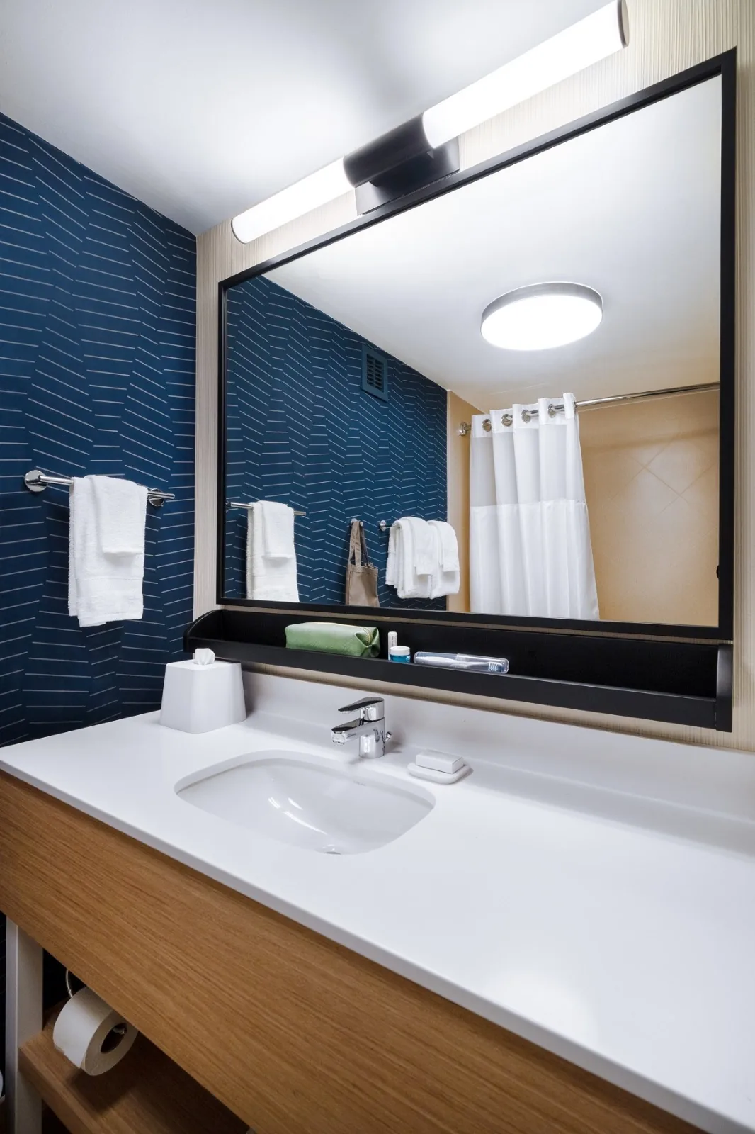 A guest bathroom vanity at Spark by Hilton, navy patterned wallcovering above a white countertop with a black framed mirror